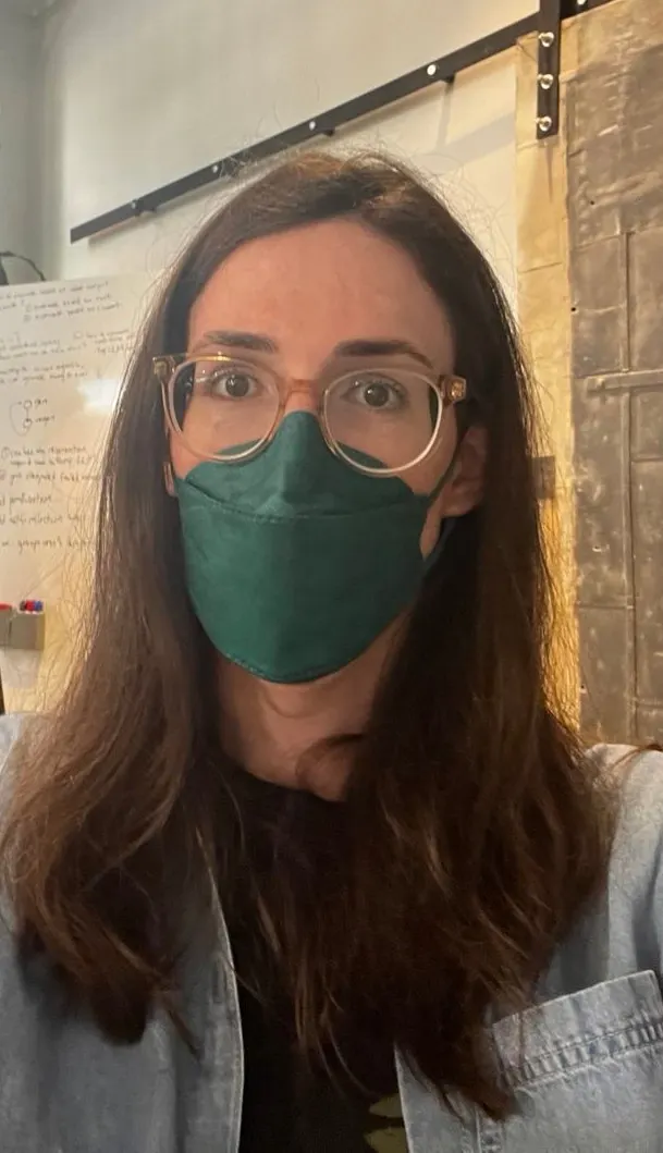 A person with long brown hair, clear glasses, and a green face mask looks directly at the camera. They wear a denim jacket, standing in a room with a whiteboard in the background.
