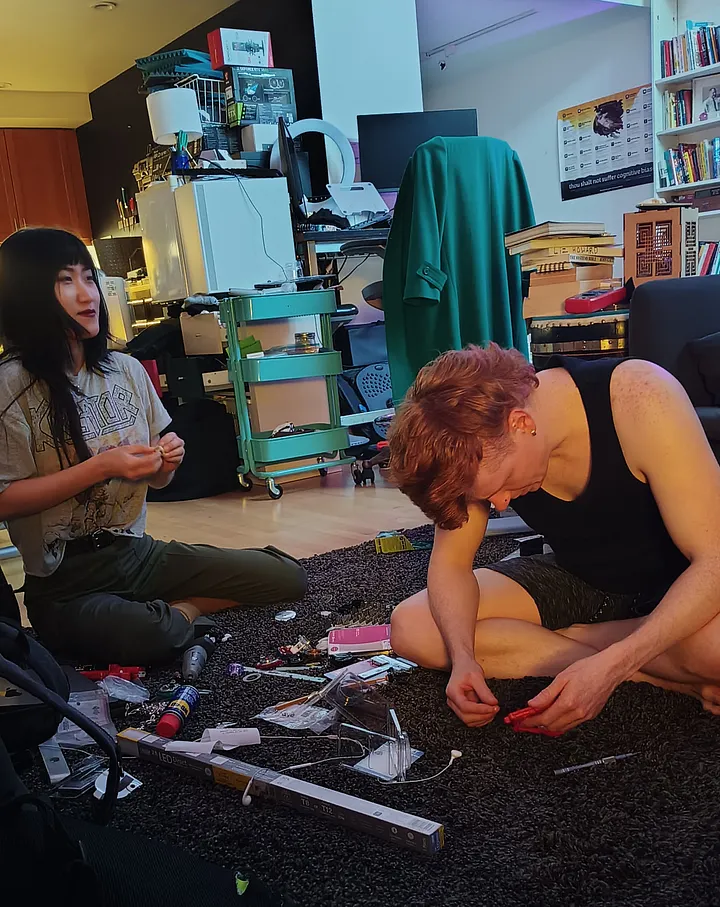 Two people sit cross-legged on a dark rug, surrounded by small parts and tools as they make modifications to a Beyblade and its launcher.