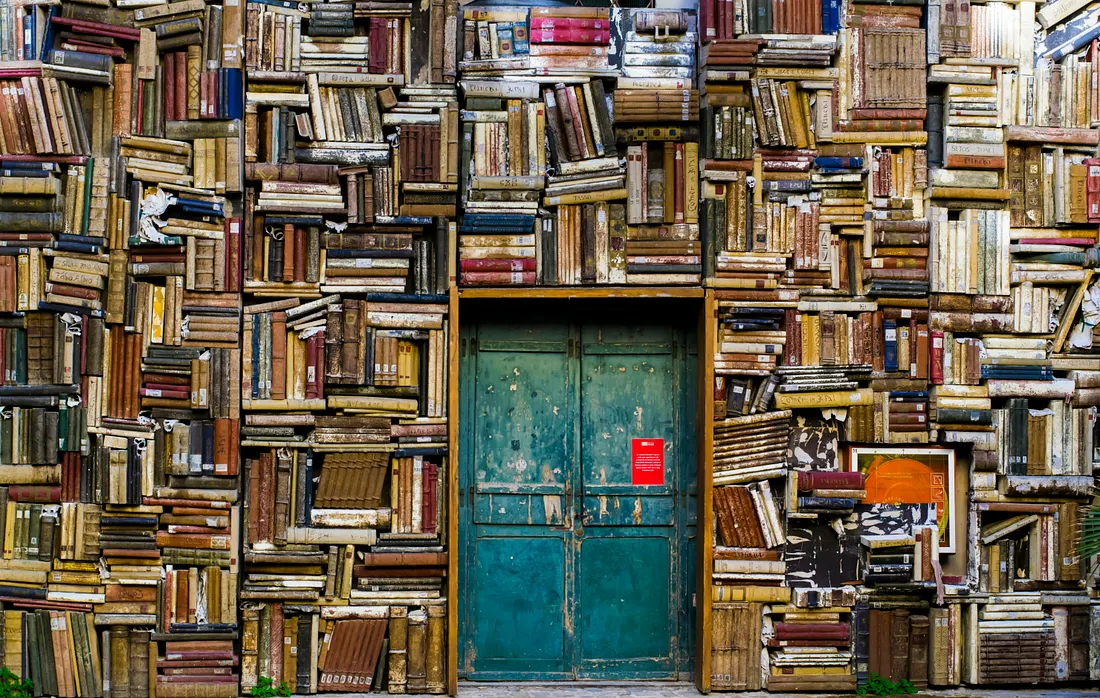 A weathered, teal double door is set into a wall made entirely of old, tightly stacked hardcover books.