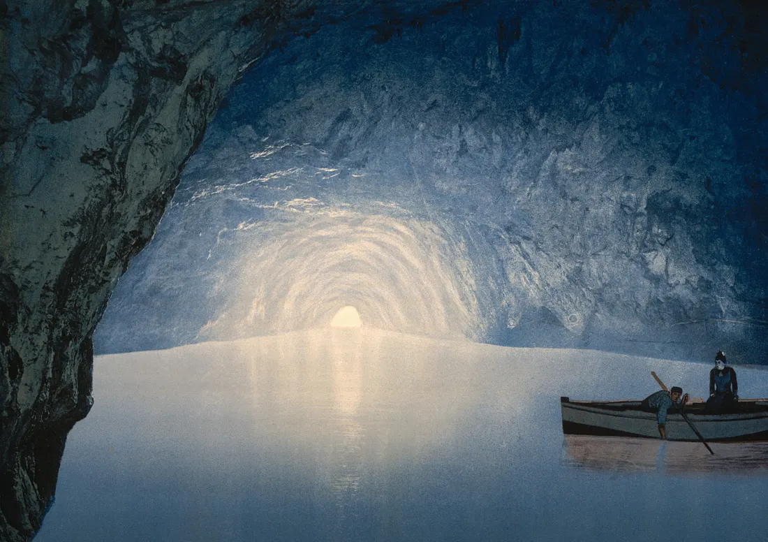 An early photochrom print shows the luminous blue interior of the Blue Grotto in Capri, Italy, with a small rowboat and figures floating towards the distant, bright cave entrance.