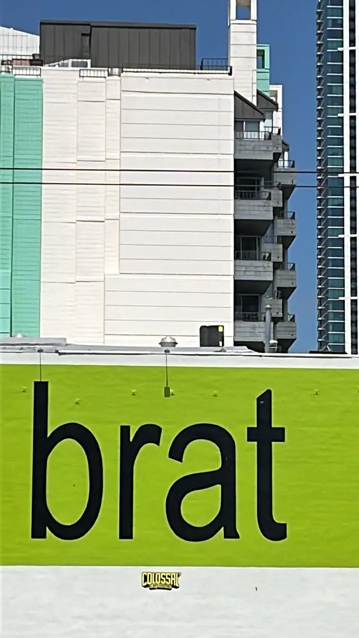 The word "brat" is written in black, lowercase text on a large, lime-green billboard. Behind the billboard, modern skyscrapers are visible under a blue sky.