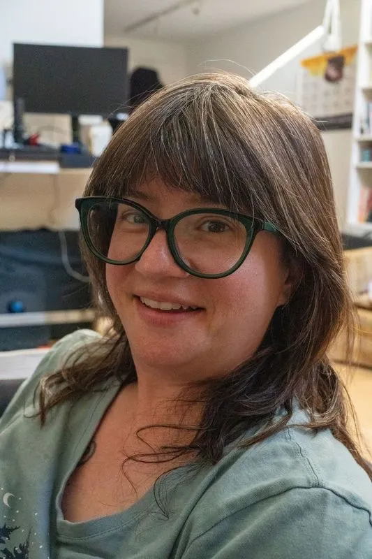 A close-up portrait of a smiling person with shoulder-length brown hair and dark, cat-eye glasses. They are in an indoor setting with a blurred office background.