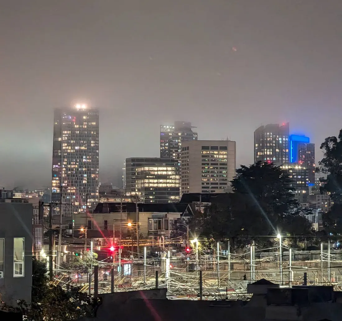 A foggy night skyline of San Francisco, looking towards the intersection of Market Street and Van Ness Avenue. a dark, fenced-off area with some low structures and visible streetlights, including glowing red traffic lights, separates the viewer from the distant city buildings. Hayes Valley is to the left and Civic Center is to the right. In the background, several illuminated skyscrapers tower above, their upper floors partially obscured by the dense fog and low clouds, with one building distinguished by its blue exterior lighting.