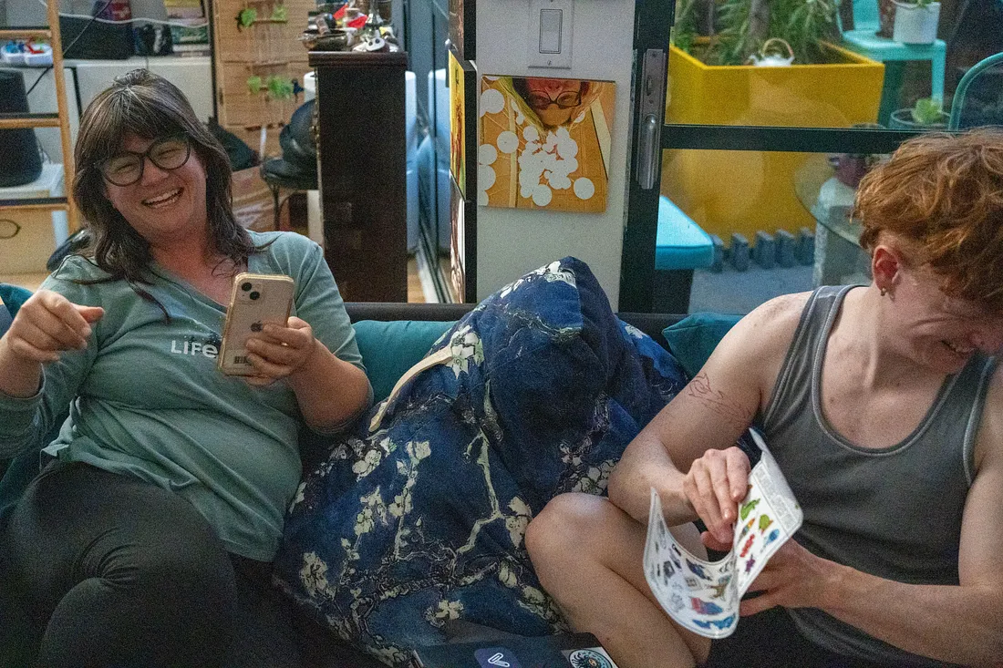 Two people sit close together on a couch, laughing. The person on the left wears glasses, holds a phone, and points forward, while the person on the right looks down at a paper booklet in their hands.