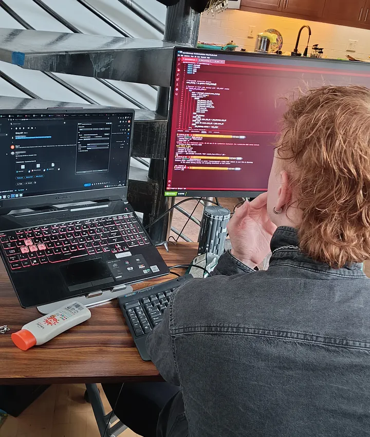 A person with shoulder-length wavy hair sits at a desk, viewed from behind, looking at two monitors. One large vertical monitor displays red text on a dark background, while a laptop on a stand shows a dark-mode application.