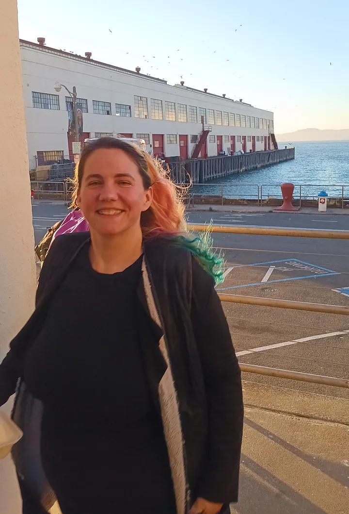 A smiling person with pink and teal hair stands outdoors in the sun, with a large white pier building, a parking lot, and a body of water visible in the background.