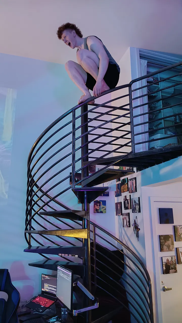 A person crouches on the metal railing at the top landing of a spiral staircase, looking down with their mouth wide open. Below, a computer desk with a glowing keyboard is visible in a room with blue and purple lighting.