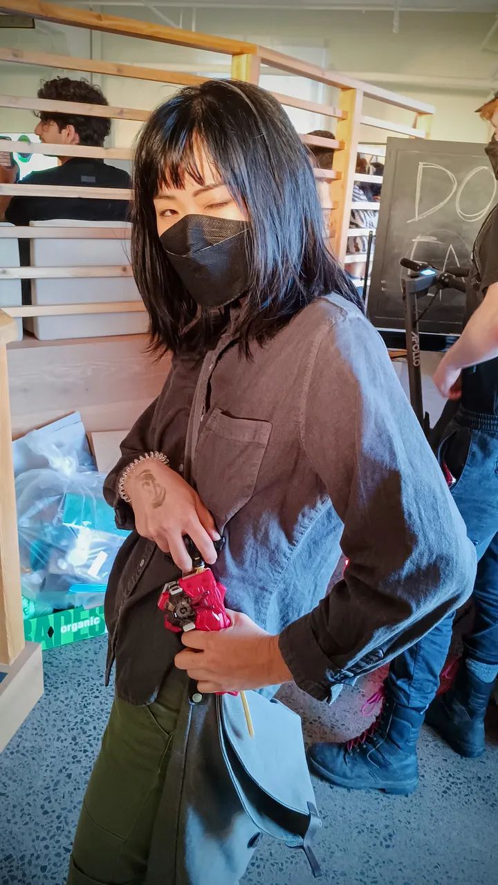 A person with shoulder-length black hair and a face mask winks at the camera while holding a red Beyblade launcher.