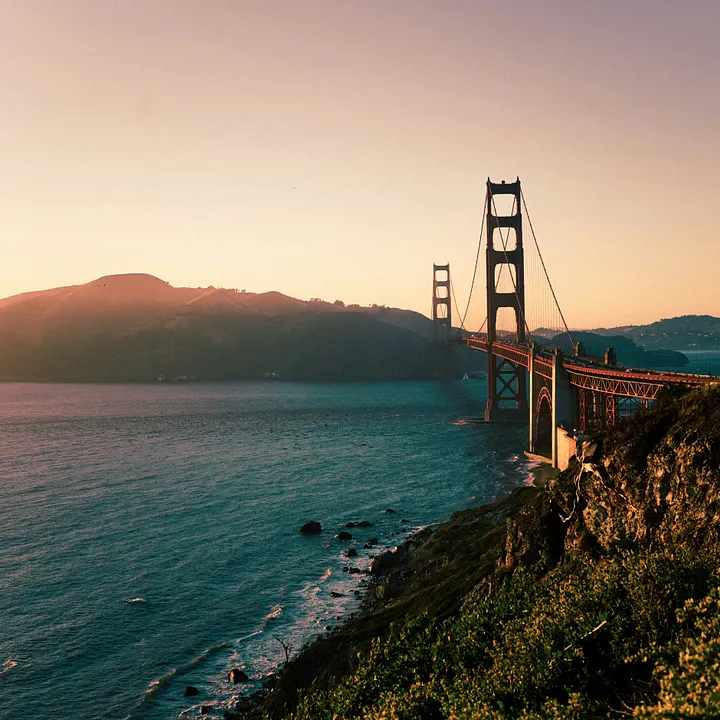 A scenic view of the Golden Gate Bridge at sunrise. The bridge's iconic red-orange towers and suspension cables stretch from the right foreground across the blue water of the bay toward rolling, sunlit hills in the background, all under a soft, warm-toned sky.