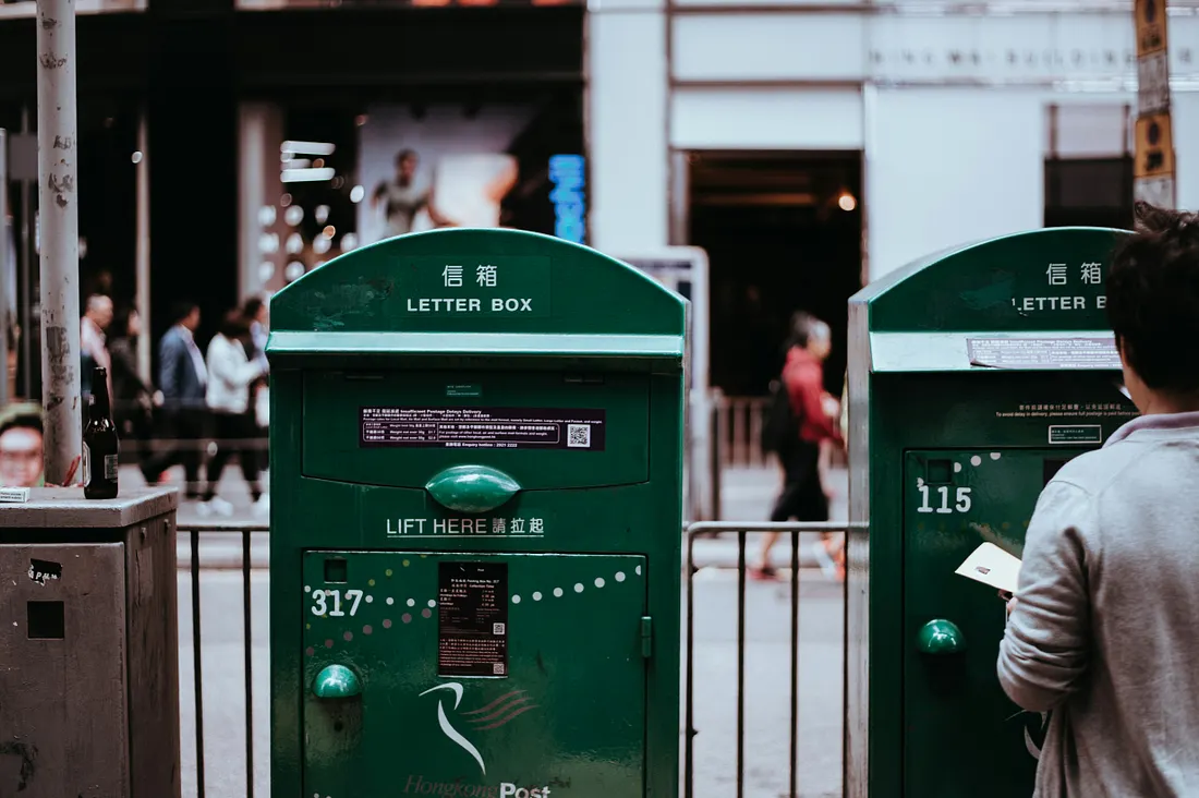 A person in a grey sweater stands holding a letter in front of two dark green Hongkong Post mailboxes on a busy city sidewalk, with pedestrians and storefronts blurred in the background.