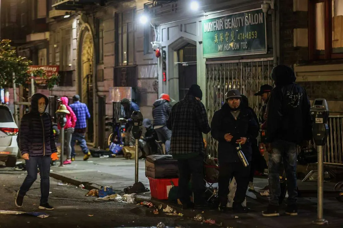 A group of people are gathered on a littered city sidewalk at night, standing and walking in front of a building with a lit "Mayfair Beauty Salon" sign.