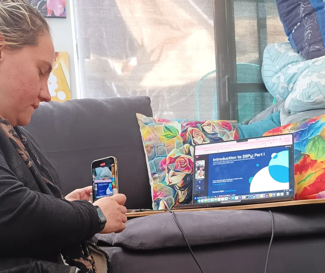 A person sits on a grey couch, simultaneously holding a smartphone in their left hand and looking at a laptop displaying a presentation titled "Introduction to DSPy: Part 1".
