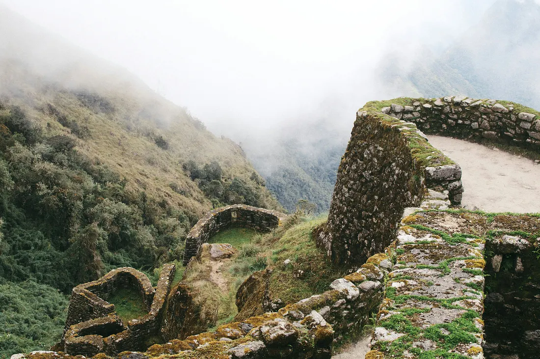 Moss-covered stone ruins, including a curved walkway and circular structures, are set into a steep, grassy mountainside shrouded in thick white fog.