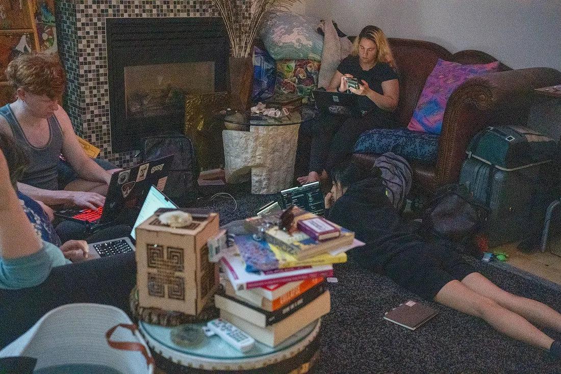 Four people are gathered in a living room, each focused on a laptop or phone. Three people sit on a couch while the other one is on the floor, surrounded by backpacks, stacks of books, and a tiled fireplace.