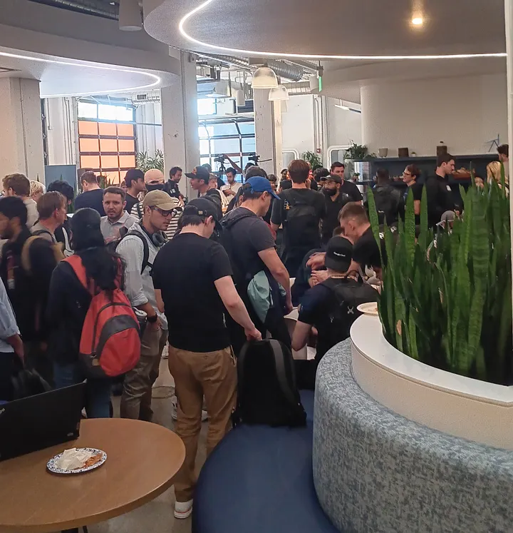 A large group of people gathers in a modern, brightly lit office space. Some individuals are standing and conversing, while others are seated at tables or on curved benches.