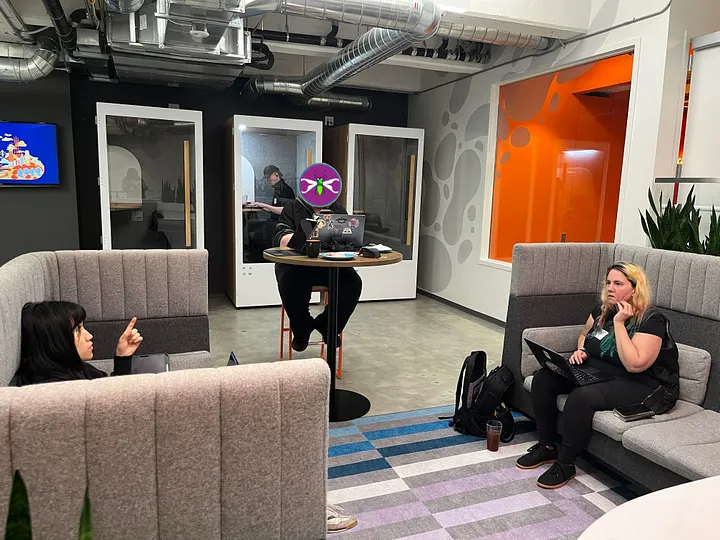 Four people are situated in a modern office lounge, with two seated in high-backed grey booths and a third at a high-top table with a laptop. In the background, white privacy pods line the wall near a bright orange accent panel, one occupied by a person in all black clothing.