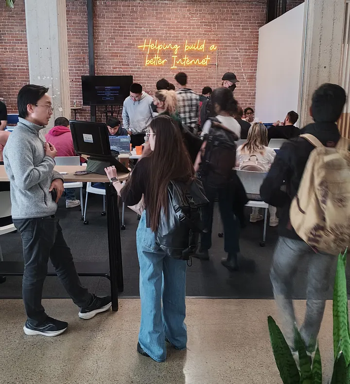 Two people stand and talk at a high-top table in a crowded event space, one facing a laptop. In the background, other attendees are gathered in front of an exposed brick wall featuring a yellow neon sign that reads, "Helping build a better Internet."