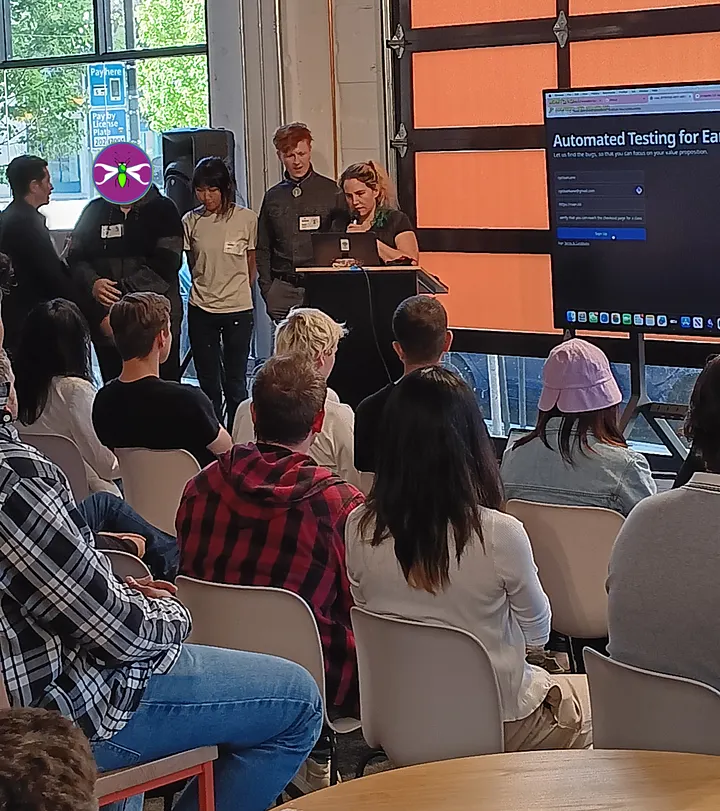 A group of people stands at the front of a room, presenting to a seated audience. A large screen behind the presenters showcases the teams early stage startup QA testing agent.