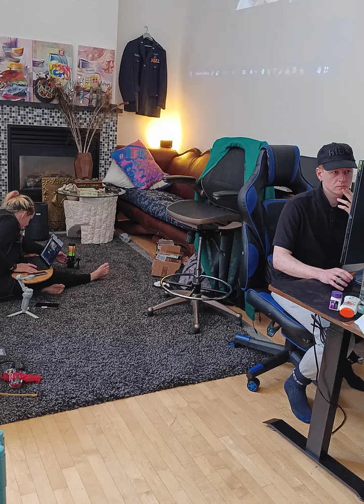 In a cluttered living room, a person in a baseball cap sits at a desk in a gaming chair, while another person sits on the floor to their left using a laptop.