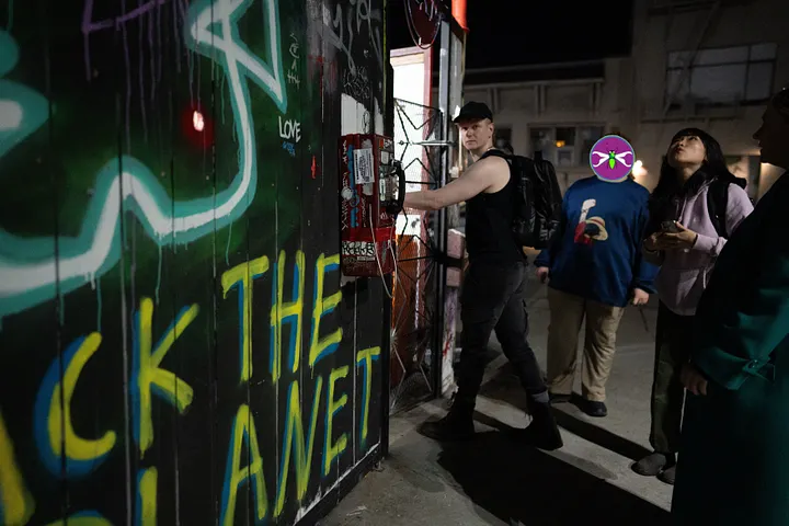 A small group gathers at the graffiti-covered entrance to Noisebridge hackerspace at night.