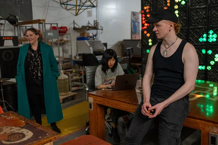 Three people are in a workshop; one person sits on the edge of a large wooden table, another uses a laptop at the same table, and a third stands smiling in a long teal coat. In the background, a large grid of glowing green, red, and orange lights is mounted on the wall.