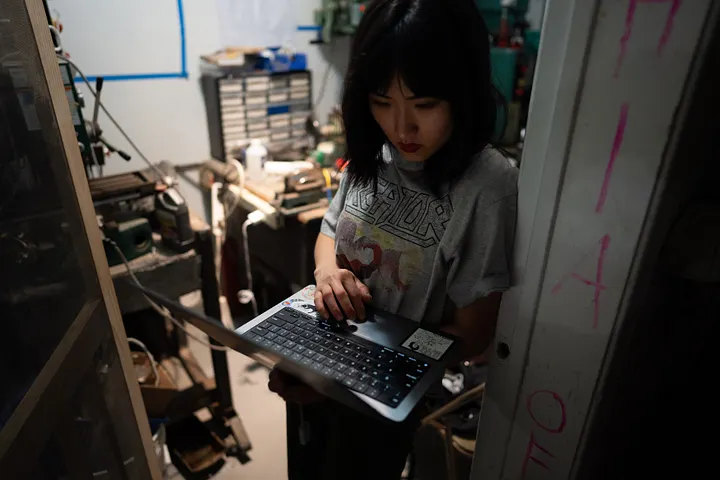 A person in a grey graphic t-shirt stands in a cluttered workshop, looking down as they hold and type on a silver laptop.