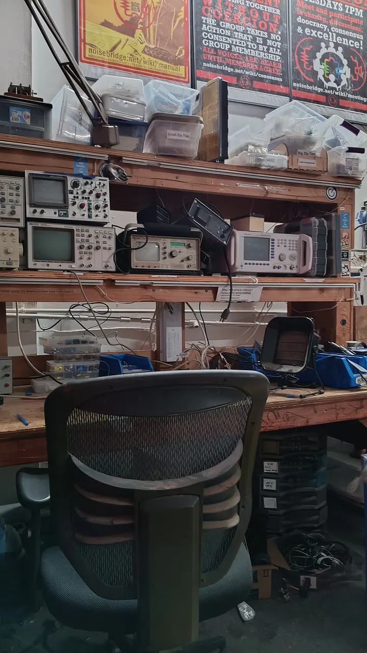 An empty, mesh-backed office chair faces a cluttered electronics workbench at Noisebridge. The wooden shelves above the bench are packed with various pieces of test equipment, including multiple oscilloscopes, power supplies, and plastic storage bins.