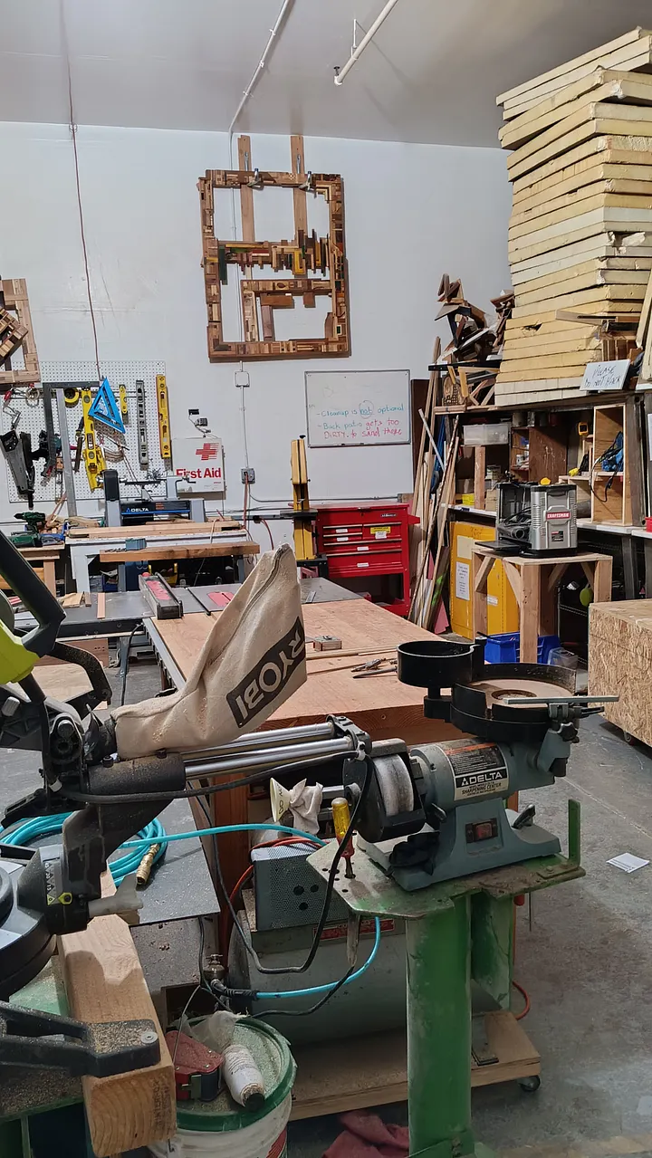 A cluttered woodworking shop filled with various power tools, including a miter saw and a bench grinder in the foreground, with workbenches and lumber stacked against the back walls.