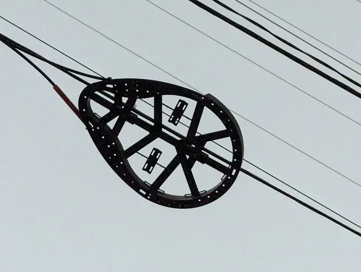 A black, oval optical fiber snowshoe holding coiled cables, seen from below against a grey sky.