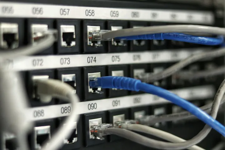 A close-up of a black, numbered network patch panel. Many blue and grey Ethernet cables are plugged into various ports.