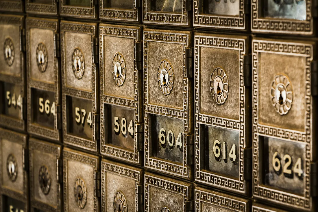 A close-up, angled shot of a wall of vintage, bronze-colored P.O. boxes, each with a numbered window, a keyhole, and intricate decorative trim.