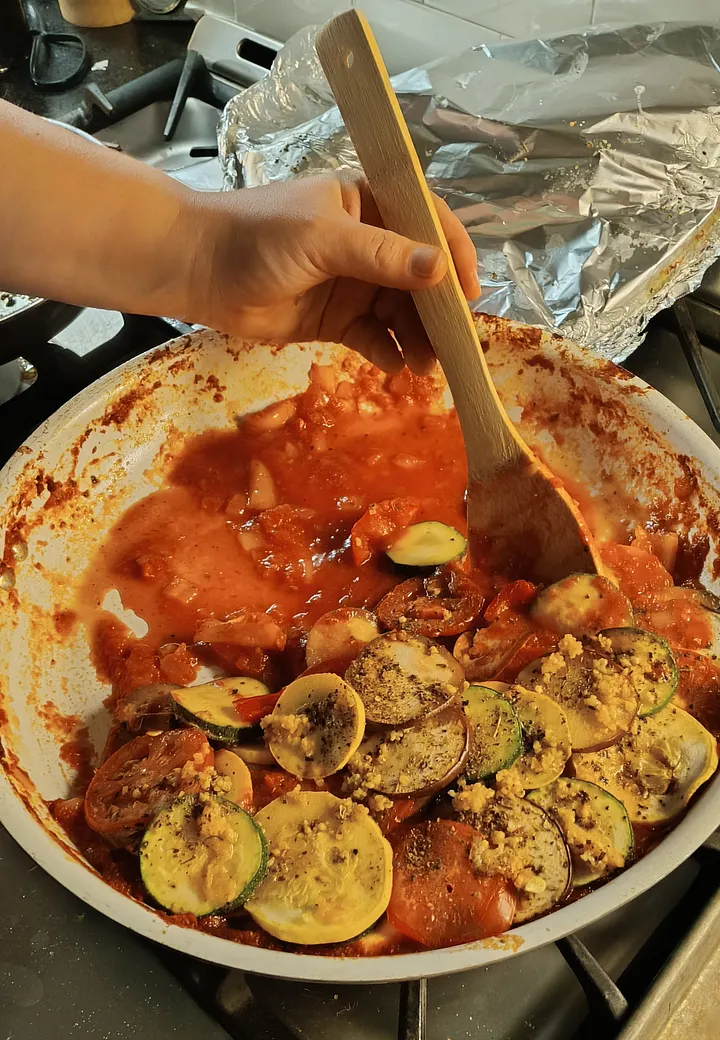 A person serves ratatouille from a white dish, using a wooden spoon to mix tomato sauce with a layer of spiraled, sliced eggplant, zucchini, yellow squash, and tomato.