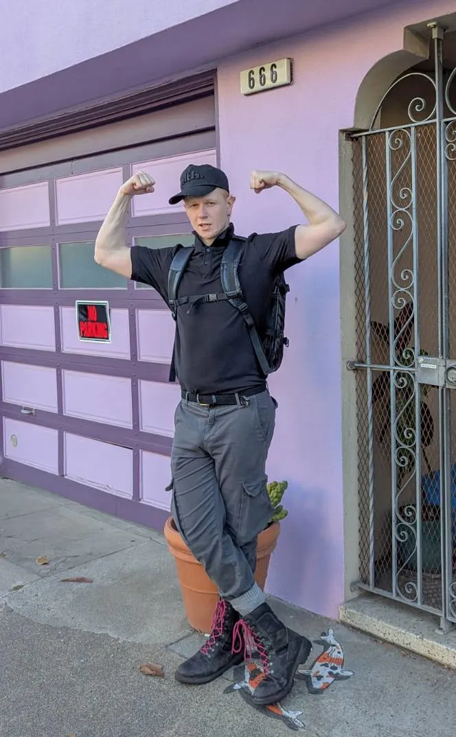 A person wearing a backpack, backward cap, and boots stands on a sidewalk, flexing both biceps. They are posing in front of a light purple building with a purple garage door and a house number plaque reading "666".