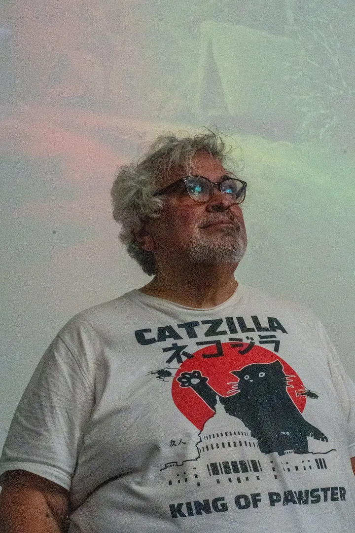 A person with gray, wavy hair and glasses looks up and to the right against a multicolor background. They are wearing a white t-shirt featuring a "Catzilla" graphic, which depicts a giant black cat climbing the U.S. Capitol building.