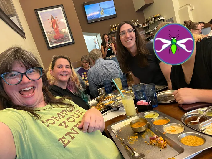 A person in a green t-shirt takes a smiling group selfie at a restaurant table covered in metal thali plates of Indian food. Several other people are gathered around the table, looking at the camera.