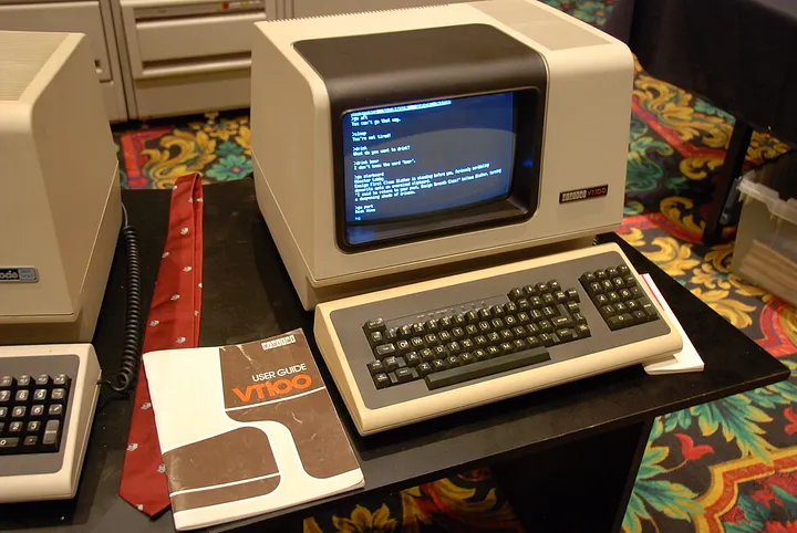 A vintage, beige Digital Equipment Corporation (DEC) VT100 terminal, with its CRT monitor and attached keyboard, is displayed on a table. The screen is illuminated with white text on a black background, and a "VT100 User Guide" rests to the left of it, lying on the desk.